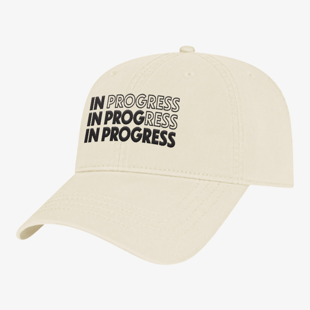 Beige cap with 'IN PROGRESS IN PROGRESS IN PROGRESS' text on a white background