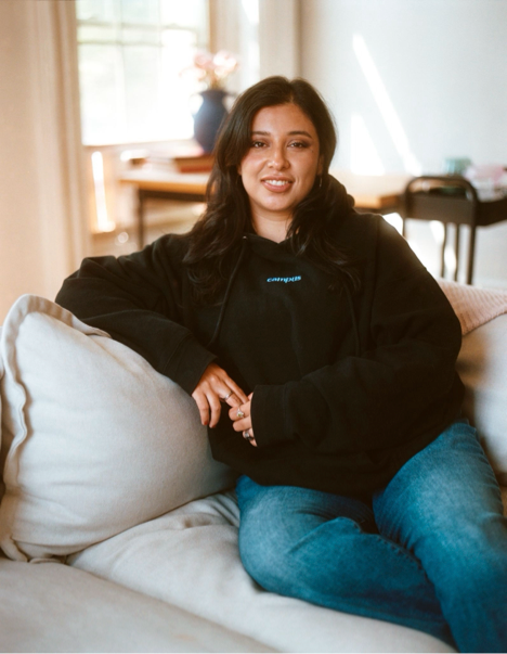Woman wearing a black hoodie with a logo, sitting on a couch in a home setting.
